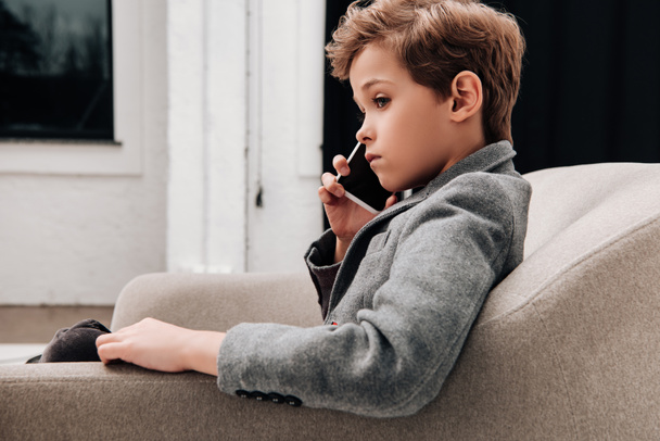 stylish little boy sitting in armchair and talking by phone - Foto, Imagen