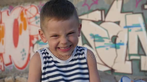 portret zabawny mały chłopiec w pasiastej koszuli, kto krzyczy, grymasy i uśmiechał się, patrząc na kamery na tle ściany z graffiti - Materiał filmowy, wideo