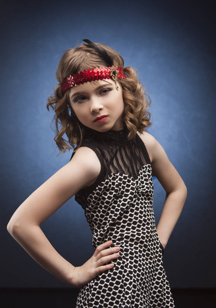 Beautiful little fashion model walking on catwalk in studio on background lighted with blue. Aggressove look, red lips and dressed in the style of a 20's Great Gatsby on party - Фото, изображение
