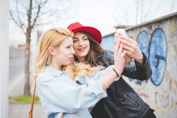 若い美しい金髪とブルネットの女の子がスマート フォンで selfie を取って街で楽しんで  - 写真・画像