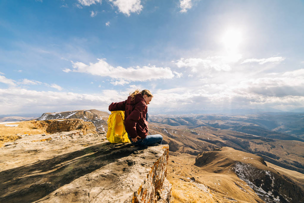 active young girl sitting on the edge of the cliff, with a yellow backpack, enjoying the mountain air - Photo, Image