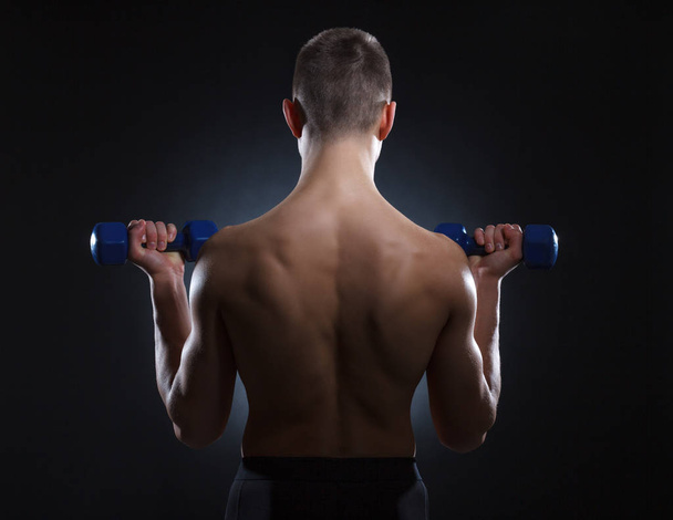 Muscular young man with dumbbells from behind on a dark background. - Photo, Image