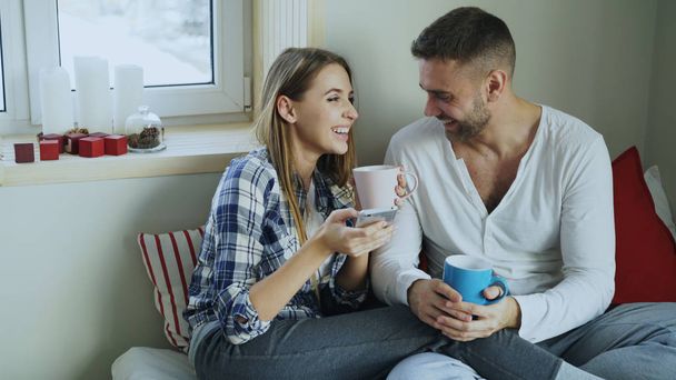 Happy young couple talking and laughing while sitting in on bed and drink coffe in the morning at home - Φωτογραφία, εικόνα
