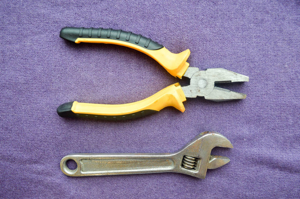 Metal, iron, shiny, adjustable wrench and pliers with rubber black-and-yellow handles against the background of purple cloth. Bench tools. - Photo, Image