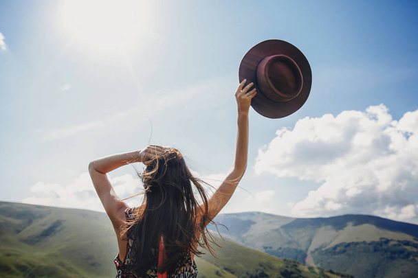 stylish traveler woman looking at mountains. hipster girl on top of mountain, raising hands and holding hat in sky. space for text. atmospheric epic moment. wanderlust and travel concept - Foto, imagen