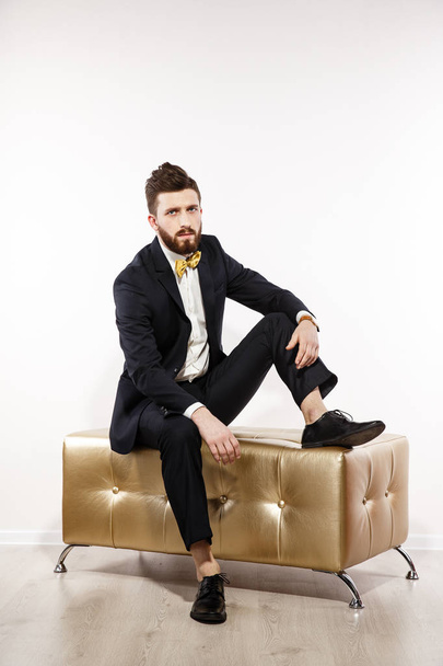 Elegant young handsome man in black classic costume. Studio fashion portrait. Sit on gold sofa. - Фото, зображення