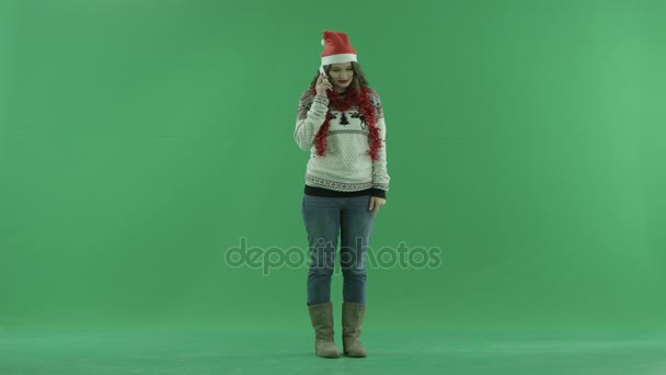 Mujer joven emocionada en sombrero de Navidad hablando sobre su teléfono inteligente mientras está de pie, croma clave en el fondo
 - Metraje, vídeo