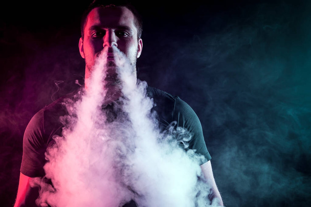 Νέοι κομψό αρσενικό καπνιστής σε ένα μαύρο T-shirt εκπνέει μέσα ένα μεγάλο σύννεφο καπνού από vape πάνω σε μαύρο φόντο απομονωμένες - Φωτογραφία, εικόνα