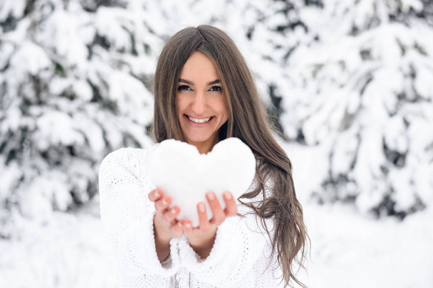 Attractive young woman in a white jumper keeps the heart out of the snow in the winter forest and smiles in the winter forest - Photo, Image