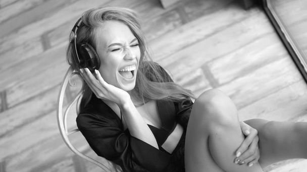 Ακρόαση μουσικής μέσω ακουστικών στο σπίτι και τραγουδούν το πρωί νεαρή γυναίκα. Φοράει εσώρουχα μαύρη δαντέλα και μετάξι ρόμπα - Φωτογραφία, εικόνα
