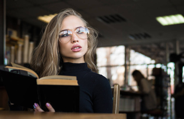 young woman student in eyeglasses, bored to read book in a library - Foto, Bild