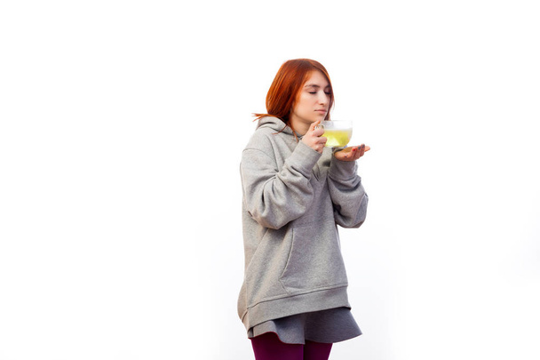Young red-haired woman in sports sweatshirt gets sick and drinks from large transparent mug antiviral medicine with vitamin with thermometer on white isolated background - Fotografie, Obrázek