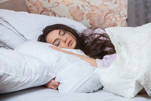 Девушка спит в белой кровати дома. Молодая женщина спит в сонной одежде на белом белье в постели дома, вид сверху
. - Фото, изображение