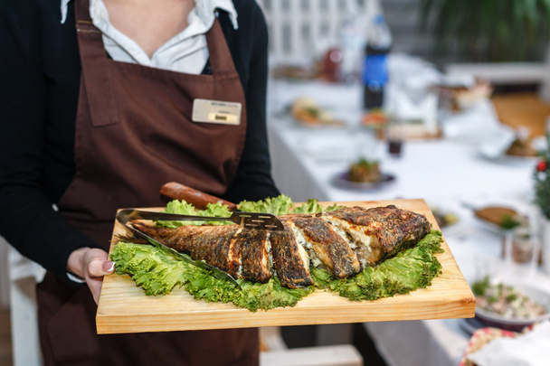 Kellnerin in schwarzer Uniform hält einen Holzteller mit gebackenem Fisch in der Hand - Foto, Bild