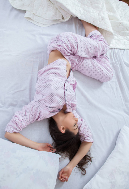 Девушка спит в белой кровати дома. Молодая женщина спит в сонной одежде на белом белье в постели дома, вид сверху
. - Фото, изображение