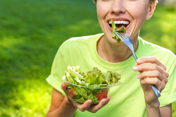 Kobieta jedzenie zdrowe sałatki z plastikowym pojemniku na zielonej trawie z latające włosy w słoneczny wiosenny dzień. podświetlenie - Zdjęcie, obraz