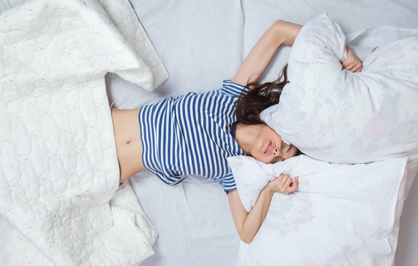 Молодая красивая женщина просыпается в светлой комнате. Расслабленная женщина лежит в постели в сонной одежде
. - Фото, изображение