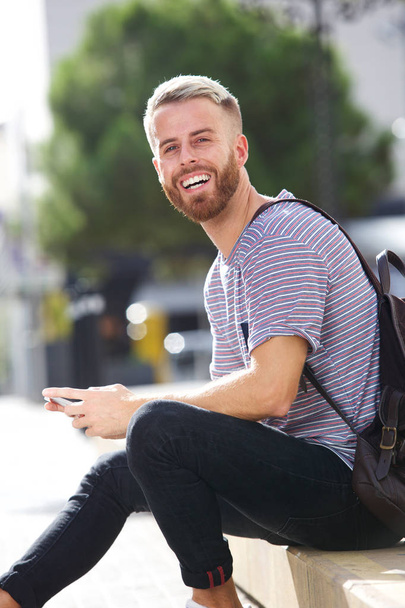 Retrato de un estudiante universitario sonriente sentado afuera con teléfono móvil
 - Foto, imagen