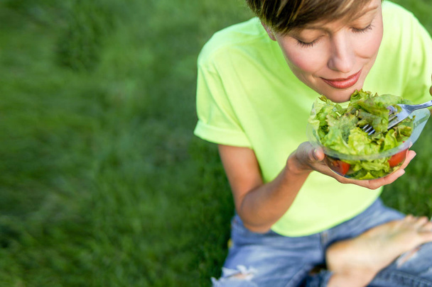 Szczupła dziewczyna jedzenia warzyw Sałatka na zewnątrz siedzi na zielonej trawie. Zdrowy styl życia, miejsce - Zdjęcie, obraz