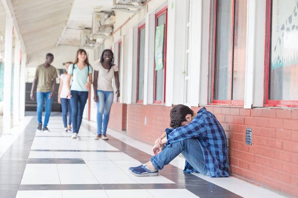School tegen pesten. Kaukasische mannelijke tiener wanhopige zitten in de hal van de school terwijl groep tieners lopen naar hem. - Foto, afbeelding