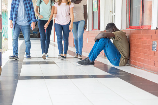 School tegen pesten. Afro Amerikaanse mannelijke tiener overstuur zitten in de hal van de school terwijl groep tieners lopen naar hem. - Foto, afbeelding