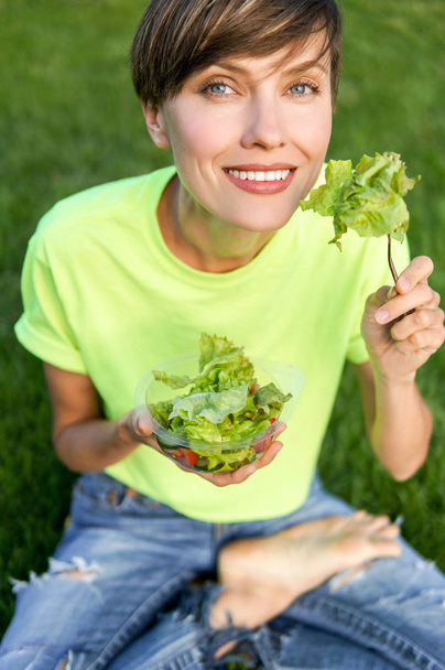 Happy smiling girl eating vegetables salad outdoors sitting on green grass. Healthy lifestyle, copy space - Photo, Image
