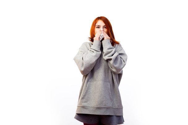 A young red-haired woman in a gray sweatshirt got a cold, is about to sneeze and covers her mouth with a white disposable napkin on a white isolated background - Foto, imagen