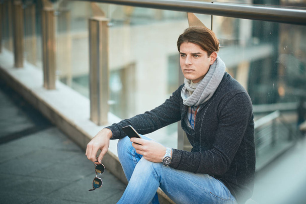 Młody mężczyzna siedzący odkryty i przy użyciu smartfona. - Zdjęcie, obraz