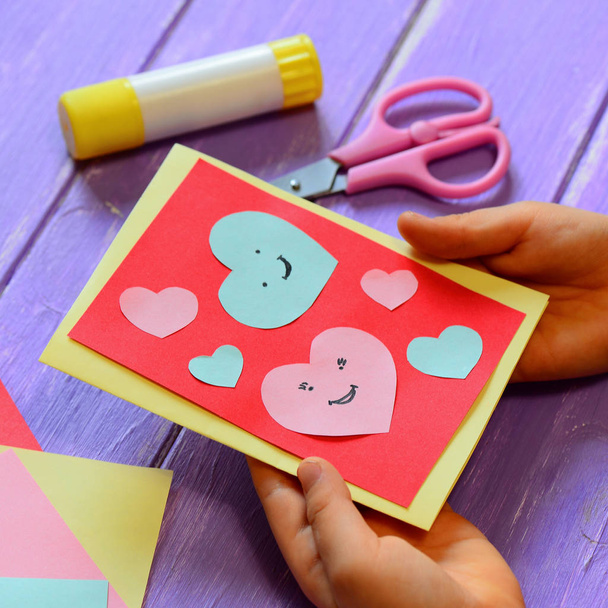 Child is holding a Valentines day greeting card in his hands. Small child is showing a Valentines day greeting card gift surprise. Cute and easy paper art for kindergarten. Paper cut out hearts. Closeup. Paper crafts for home. Easy paper crafts kids - Photo, Image