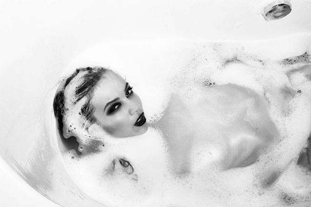 One attractive sensual sexy young woman with bright makeup and wet naked body lying in white bath in bathroom full of water and soap foam indoor on studio background, horizontal picture - Foto, Bild