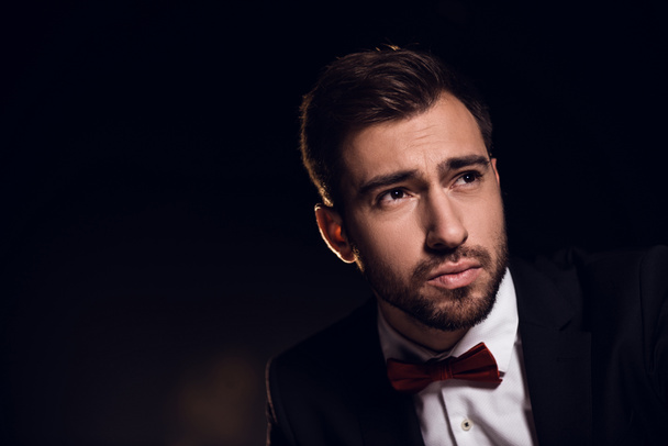 sad bearded handsome man in jacket and bow tie, isolated on black - Фото, изображение