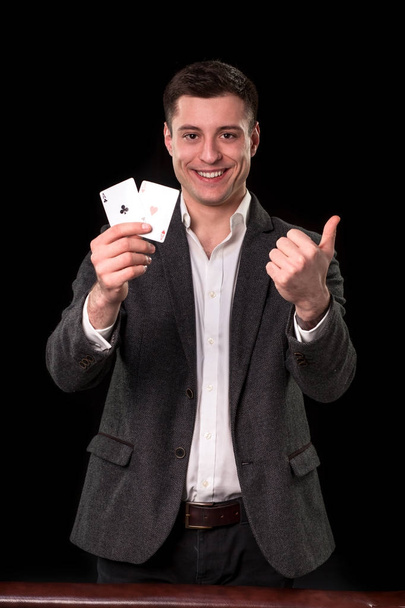 Young caucasian man wearing suit holding two aces in his hand and showing thumb up hand gesture with smile on black background. Gambling concept. Casino - Photo, image