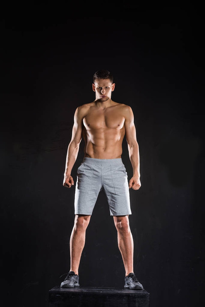 full length view of shirtless muscular sportsman standing on block and looking at camera on black - Fotografie, Obrázek