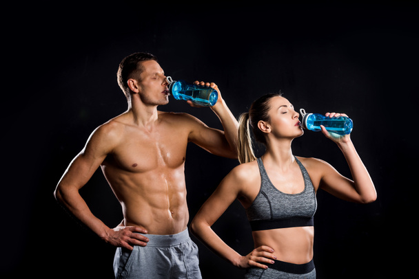 athletic young couple in sportswear drinking from bottles isolated on black - Photo, Image