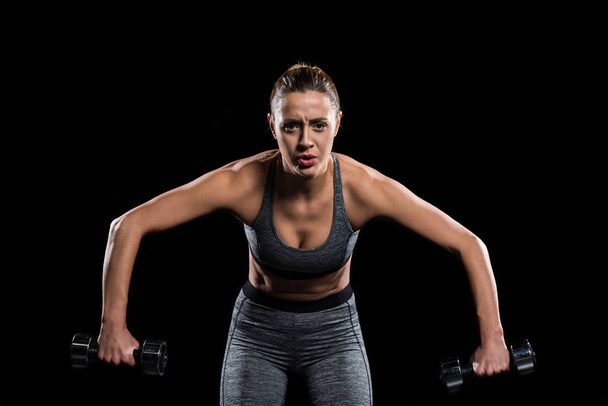 tired sporty young woman exercising with dumbbells and looking at camera isolated on black  - Photo, Image
