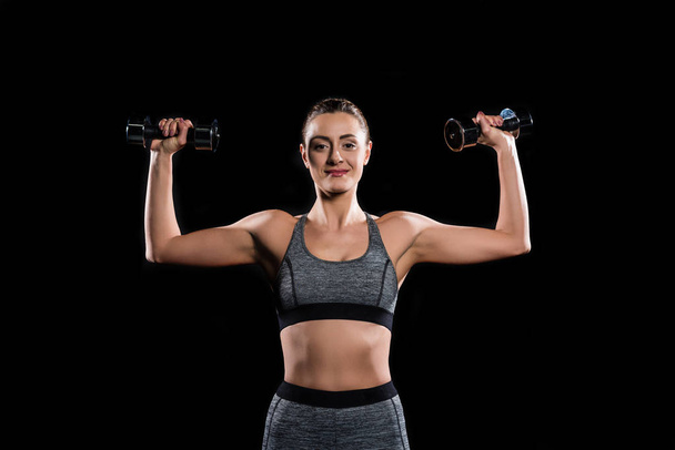 young sportswoman holding dumbbells and smiling at camera on black  - Photo, Image