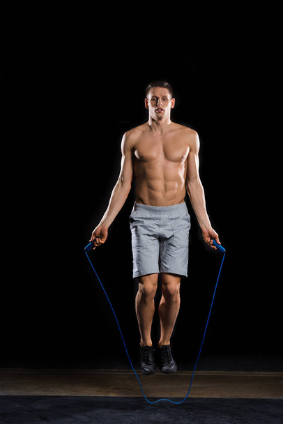 muscular shirtless sportsman jumping with skipping rope on black - Фото, изображение