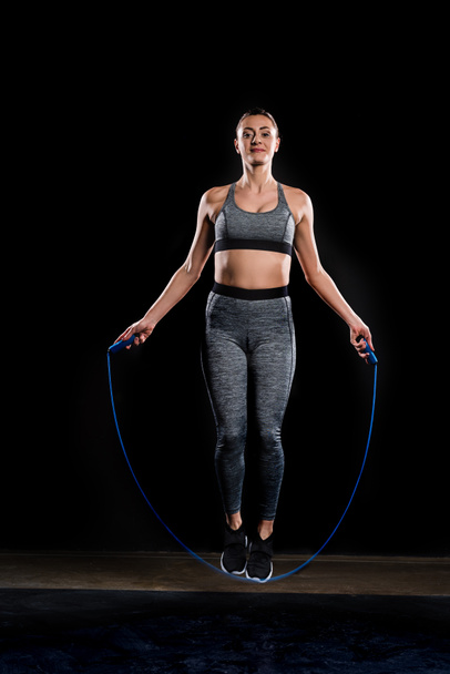 sportswoman jumping with skipping rope and looking at camera on black - Photo, Image