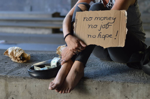 eine obdachlose reife Punkerin, die unter einer Brücke sitzt, bettelt und ein Schild hält - kein Geld, keine Arbeit, keine Hoffnung - Foto, Bild