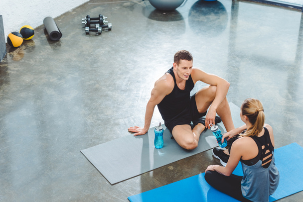 vista de ángulo alto de deportista y deportista con botellas de agua sentados en colchonetas de yoga y mirándose en el gimnasio
 - Foto, imagen