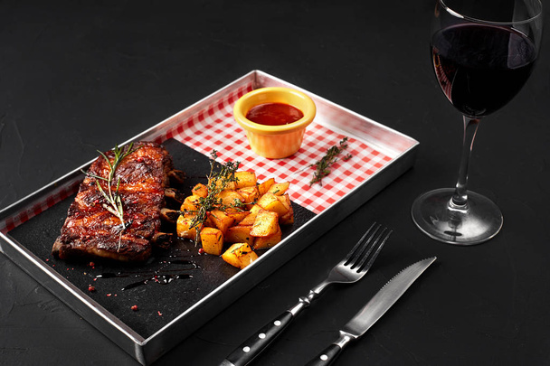 Delicious barbecued ribs seasoned with a sauce, potatoes and served with herbs on a metal tray, on black background - 写真・画像