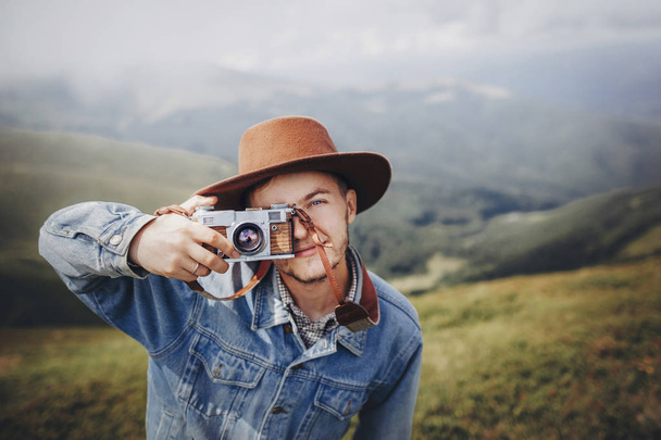 стильний чоловік мандрівник у капелюсі з фотоапаратом, посміхаючись на вершині гір. стильний хіпстер робить фотографію. атмосферний момент
. - Фото, зображення