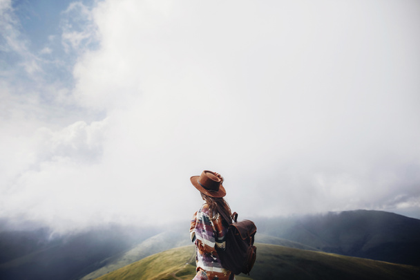 wanderlust and travel concept. girl traveler in hat with backpack looking at clouds in mountains. stylish hipster woman exploring on top of mountain. space for text. atmospheric moment - 写真・画像