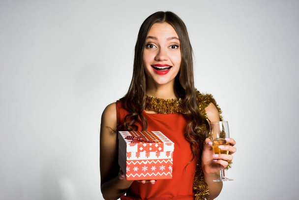 happy young girl with tinsel on the neck celebrating the new year, holding a glass of champagne and a gift - Foto, Imagen