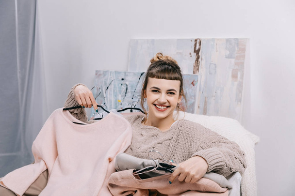 улыбающаяся девушка сидит на кресле с одеждой на вешалках и смотрит в камеру
 - Фото, изображение