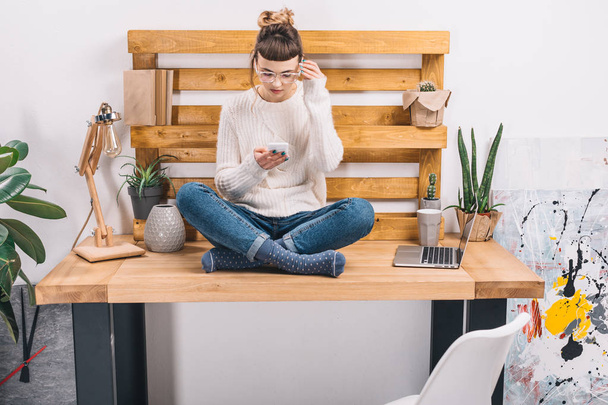 girl sitting on table in office and looking at smartphone - Photo, Image