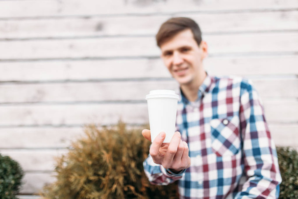 A man holding a white paper Cup, a handsome guy, in a cellular shirt, on the background of a wooden wall - Foto, imagen