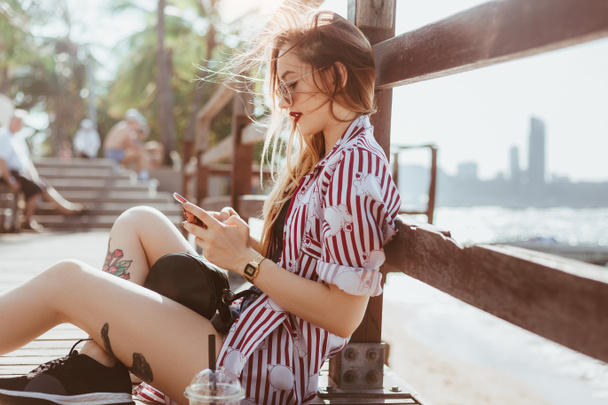 piękna młoda kobieta przy użyciu smartfona siedząc na molo na plaży - Zdjęcie, obraz
