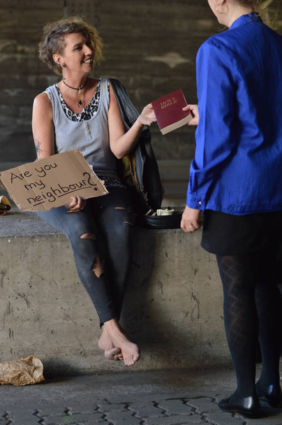 eine Punk-Frau sitzt unter einer Brücke mit einer Flasche Alkohol neben einem Schild und bekommt eine Bibel von einer Geschäftsfrau - Foto, Bild