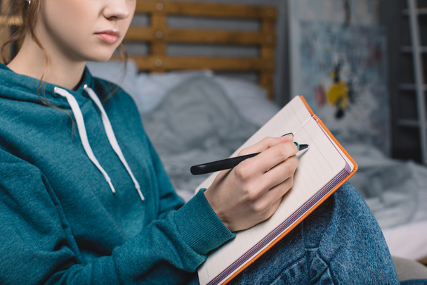 bijgesneden afbeelding van meisje zittend op fauteuil in de slaapkamer en het schrijven van iets op notebook - Foto, afbeelding